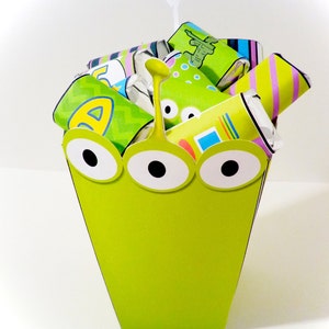 May include: A lime green alien-themed container filled with wrapped candies. The container has three large eyes and a yellow antenna. The candies are wrapped in various colorful patterns and designs, creating a playful and festive appearance.