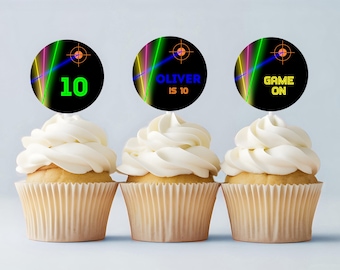 May include: Three cupcakes with white frosting and decorative toppers. The toppers are black circles with neon designs and text that reads "10", "Oliver is 10", and "Game On". The cupcakes are in beige paper cups.