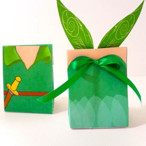 Puede incluir: Dos cajas de regalo de papel verde y marrón. Una caja presenta una imagen de dibujos animados de Peter Pan con una espada y un cinturón marrón. La otra caja tiene alas de hada verdes en la parte superior y una cinta verde atada alrededor.