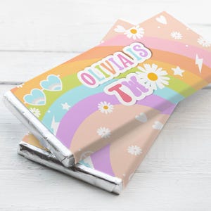 May include: Two chocolate bars with foil wrapping and rainbow-themed paper. The wrappers display daisies, hearts, and the text "Olivia's 2nd". The bars are stacked on a white wooden surface.