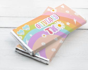 May include: Two chocolate bars wrapped in foil and colorful paper. The wrappers feature a rainbow design with daisies, hearts, and the text "Olivia's 2nd". The bars are stacked on a white wooden surface.