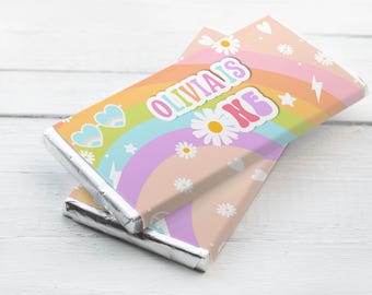 May include: Two chocolate bars wrapped in silver foil, stacked on a white wooden surface. The wrappers feature a rainbow design with daisies, hearts, stars, and the text "OLIVIA'S KE" in a colorful, retro font.