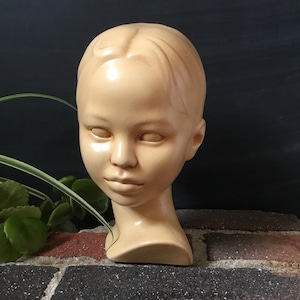 May include: A beige-colored ceramic head sculpture of a young person. The sculpture has a smooth finish, with detailed facial features and a serene expression. The head is mounted on a short neck, and the background is a dark color.