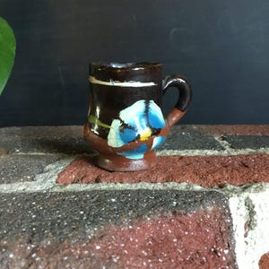 May include: Small, hand-painted ceramic mug with a dark brown base and handle. The mug features a blue floral design with yellow accents. A white stripe encircles the top rim. The mug is approximately 3 inches tall.