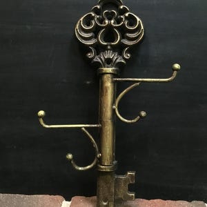 May include: An antique-style key-shaped wall hook in a bronze finish. The decorative key features ornate detailing on the head and three curved hooks for hanging items. The key is mounted on a brick surface.