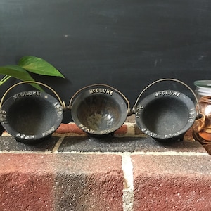 May include: Three cast iron bean pots with wire handles are arranged in a row. Each pot is black with the word "McClure" and the year 1776 or 1918 embossed on the rim. The words "Bean Soup" are also visible.