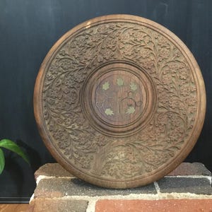 May include: A round, wooden decorative tray with intricate floral carvings. The tray has a central medallion with a leaf design and is displayed on a brick surface. The wood has a warm, brown tone.