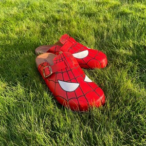 May include: Red Spider-Man clogs with a black web design and white eye shapes. The clogs have a buckle fastening. They are pictured on green grass.
