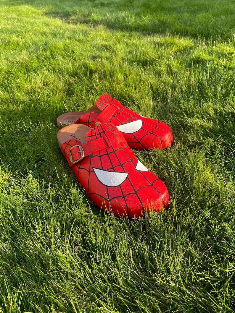 May include: Red Spider-Man clogs with a black web design and white eye shapes. The clogs have a buckle fastening. They are pictured on green grass.