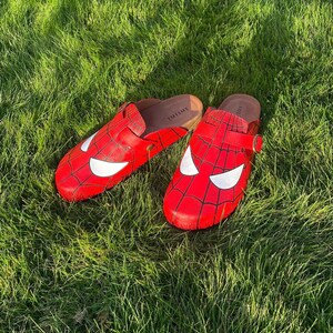 May include: Red clogs with a Spider-Man design, including a white eye mask and black web pattern. The clogs are set on green grass, offering a playful take on footwear, with a comic book theme.