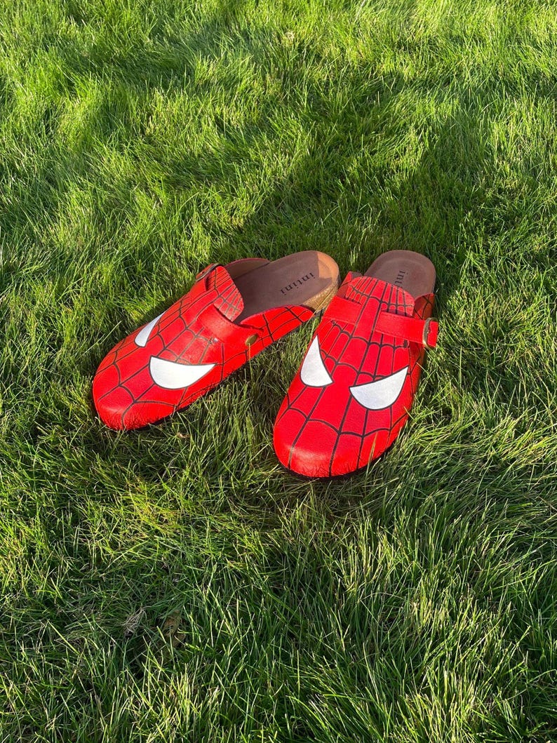 May include: Red clogs with a Spider-Man design, including a white eye mask and black web pattern. The clogs are set on green grass, offering a playful take on footwear, with a comic book theme.