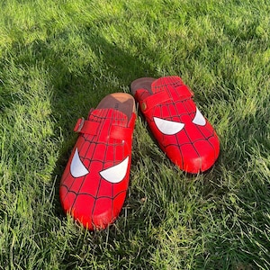 May include: Two red clogs with a Spider-Man design, showing a web pattern and white eye details. The clogs have a brown footbed and a strap with a buckle. They are placed on green grass.
