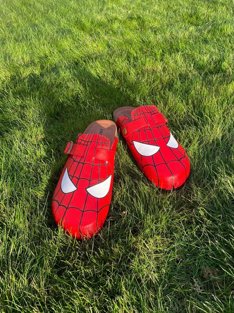 May include: Two red clogs with a Spider-Man design, showing a web pattern and white eye details. The clogs have a brown footbed and a strap with a buckle. They are placed on green grass.