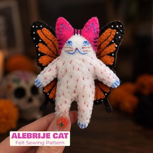 May include: A white felt cat figure with pink ears and orange monarch butterfly wings. The cat has blue embroidered eyes and whiskers, with small red stitches on its body. Text at the bottom reads ALEBRIJE CAT Felt Sewing Pattern.