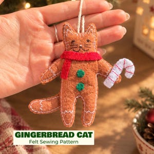 May include: A brown felt cat ornament shaped like a gingerbread cookie, featuring a red scarf, two green circular buttons on the torso, and a white and red striped candy cane held in one paw. The cat has stitched facial details.