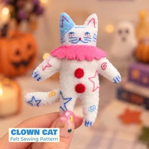 May include: A white felt cat plushie featuring a pink ruffled collar, two red circular buttons on the chest, and blue and pink embroidered star and swirl patterns. The cat has blue ears, blue embroidered eyes, and a small red nose detail.