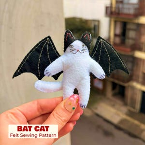 May include: A white felt cat figure with black bat wings and black ears. The cat has embroidered facial features including eyes, a nose, and whiskers. The text BAT CAT Felt Sewing Pattern is printed in red and black at the bottom of the image.