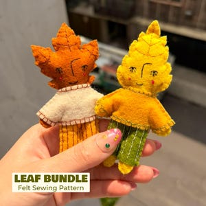 May include: Two small felt dolls shaped like autumn leaves, one orange with a white shirt and yellow striped pants, the other yellow with an orange shirt and green striped pants. Both feature embroidered facial details and visible stitching.