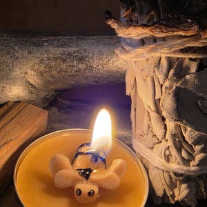 Puede incluir: Una vela encendida con forma de tortuga en un recipiente de metal. La vela está rodeada de cera amarilla y tiene un diseño azul y negro. Un manojo de salvia blanca y un trozo de madera están en el fondo.