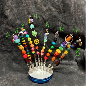 May include: A collection of eight beaded cocktail picks with various colorful beads and charms. The picks are held in a small, blue-rimmed container. Each pick features a unique design, including peace signs, turtles, and other decorative elements.