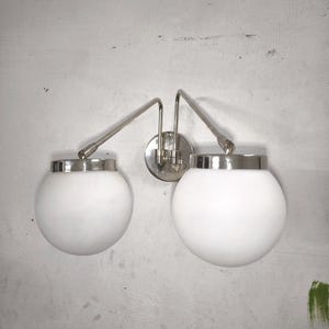 Può includere: Una lampada da parete doppia con due globi sferici in vetro bianco fissati a una piastra di montaggio centrale circolare tramite due bracci metallici regolabili color argento con finiture cromate lucide.