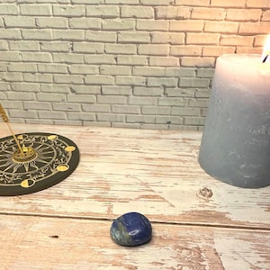 Op de afbeelding: Een blauwe en groene lapis lazuli edelsteen ligt op een licht houten oppervlak. Een zwarte wierookhouder met een gouden ontwerp en een blauwe kaars zijn ook aanwezig. De achtergrond is een witte bakstenen muur.