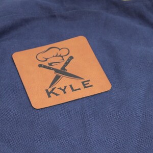 May include: Navy blue apron with a brown leather square patch featuring a chef's hat and crossed knives with the name "KYLE" printed below.