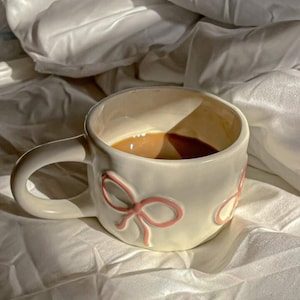 May include: A white ceramic mug with a light brown liquid inside. The mug has a handle and is decorated with pink bow designs. The mug is sitting on a white surface, possibly a bed or blanket.
