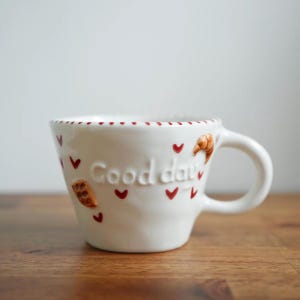 May include: A white ceramic mug with the words "Good day" embossed on the side. The mug is decorated with red hearts and small food illustrations. The rim has a dotted pattern. The mug sits on a wooden surface.