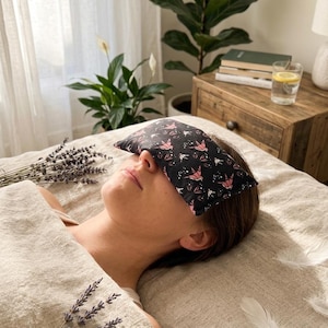 May include: A rectangular eye pillow featuring a black fabric base with a repeating pattern of pink and white origami-style birds and small white dots. The pillow is positioned across the eyes, showcasing a soft, filled construction.