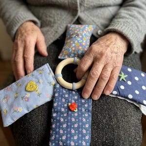 Może przedstawiać: Ręcznie robiony zestaw zabawek dla niemowląt, zawierający drewniany pierścień i cztery kawałki materiału. Kawałki materiału zdobione są wzorami kwiatowymi, kropkami i guzikiem w kształcie serca. Do zabawy sensorycznej.