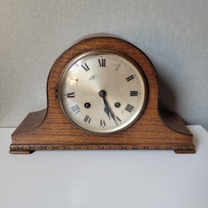 May include: A wooden mantel clock with a curved top and a rectangular base. The clock face is round with black Roman numerals and hands. The clock is made of brown wood and has decorative carvings on the base.
