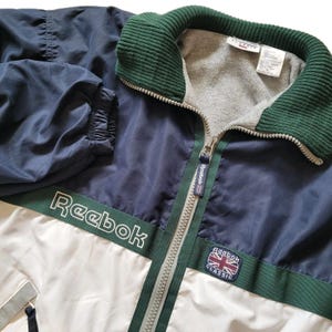 Op de afbeelding: Een vintage Reebok Classic trainingsjack in marineblauw, wit en groen. De jas heeft een volledige ritssluiting, een groene corduroy kraag en het Reebok-logo op de borst en mouw. Het heeft elastische manchetten en een wit onderste paneel.