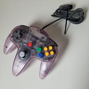 Puede incluir: Un mando de videojuegos de color morado translúcido con un joystick analógico gris central, una cruceta, un botón A rojo, un botón B verde y cuatro botones C amarillos. El mando incluye un cable negro con un conector de enchufe.