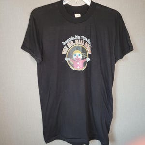May include: A black short-sleeve t-shirt featuring a graphic of a Mr. Bill character in a pink outfit inside a circular frame. The text above reads "Hey kids, it's time for..." and "THE MR. BILL SHOW" in a curved font, with "Oh nooooo kids!" to the right.