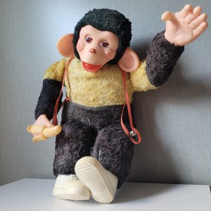 May include: A plush monkey doll with black fur on its head and limbs, a yellow torso, and dark brown trousers. It features large pink ears, a plastic face, and white shoes. The monkey holds a small plastic banana and wears orange braces.