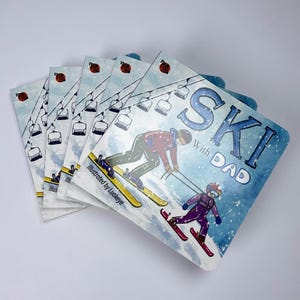 May include: A stack of square coasters with a winter sports theme. The coasters feature illustrations of skiers, ski lifts, and the words "SKI with DAD." The illustrations are in shades of blue, red, and yellow.