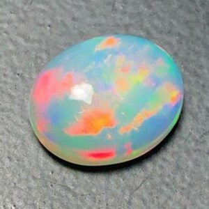 May include: Oval-shaped opal gemstone with a vibrant play of color. The stone displays a mix of red, orange, yellow, green, and blue hues. The opal has a smooth, polished surface and is set against a dark background.