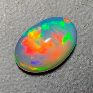 May include: A polished, oval-shaped opal gemstone. The opal displays a vibrant play-of-color, with flashes of red, orange, green, and blue hues. The stone is set against a gray surface, highlighting its iridescent qualities.