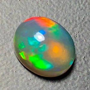 May include: A polished, oval-shaped opal gemstone. The opal displays a play of color, with flashes of red, orange, green, and blue within a translucent, gray body. The gemstone is set against a dark, textured surface.