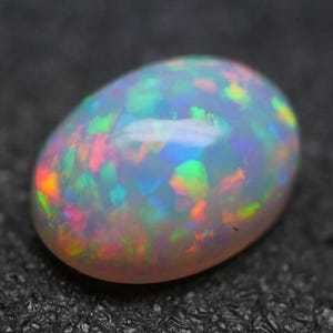 May include: Oval-shaped opal gemstone with a milky white base and vibrant flashes of color. The opal displays a play-of-color effect, with iridescent hues of blue, green, yellow, and red. The gemstone is set against a dark background.