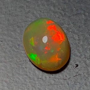 May include: Oval-shaped opal gemstone with a mix of colors. The opal features vibrant flashes of red, green, and orange, with a white spot. The gemstone is set against a gray surface.