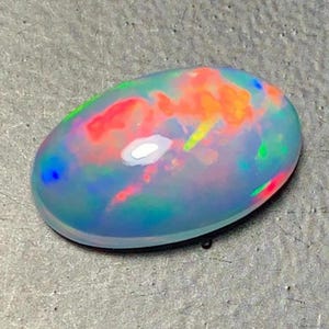 May include: Oval-shaped opal gemstone with a vibrant display of colors. The opal features a translucent base with internal flashes of red, orange, yellow, green, and blue. The stone is set against a gray surface.