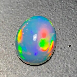 May include: A polished oval opal gemstone. The opal displays a milky white base color with vibrant flashes of red, orange, green, and blue, creating a play-of-color effect. The gemstone is set against a gray surface.