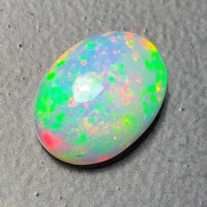 May include: A polished, oval-shaped opal gemstone. The opal displays a milky white base color with vibrant flashes of green, red, and yellow throughout. The gemstone is smooth and reflective, showcasing a play of color.