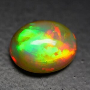 May include: A vibrant, oval-shaped opal gemstone. The stone displays a captivating play of color, with flashes of green, red, and yellow hues. The polished surface reflects light, creating a mesmerizing effect. The gemstone is set against a dark background.