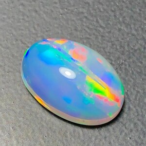 May include: An oval-shaped opal gemstone with a polished surface. The stone displays a play of color, with flashes of blue, green, orange, and red. The opal is set against a gray background.