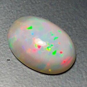 May include: A polished, oval-shaped opal gemstone. The opal has a milky white base color with flashes of green, red, and blue iridescence. The stone is smooth and appears to be well-cut, showcasing its internal play of color.