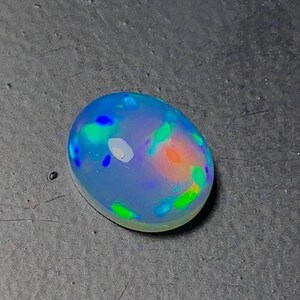 May include: An oval-shaped opal gemstone with a translucent, milky base. The stone displays vibrant flashes of color, including blue, green, orange, and yellow, creating a play-of-color effect. The opal is set against a dark gray surface.