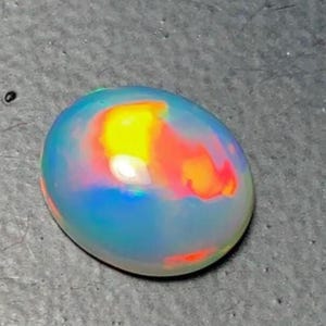 May include: A polished, oval-shaped opal gemstone. The opal displays a vibrant play-of-color, with flashes of yellow, orange, and red within a translucent, blue-hued base. The gemstone is set against a gray surface.
